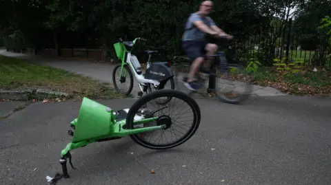 PA Media Two Lime bikes on a path, one standing and the other on its side, by a park. A cyclist is on his bike passing by in the background. 