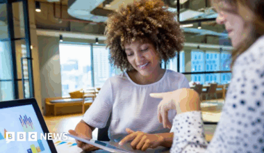 Two women sit opposite each other looking at a laptop screen and an iPad device. They are in an open-plan office with windows behind them as well as chairs and a desk.