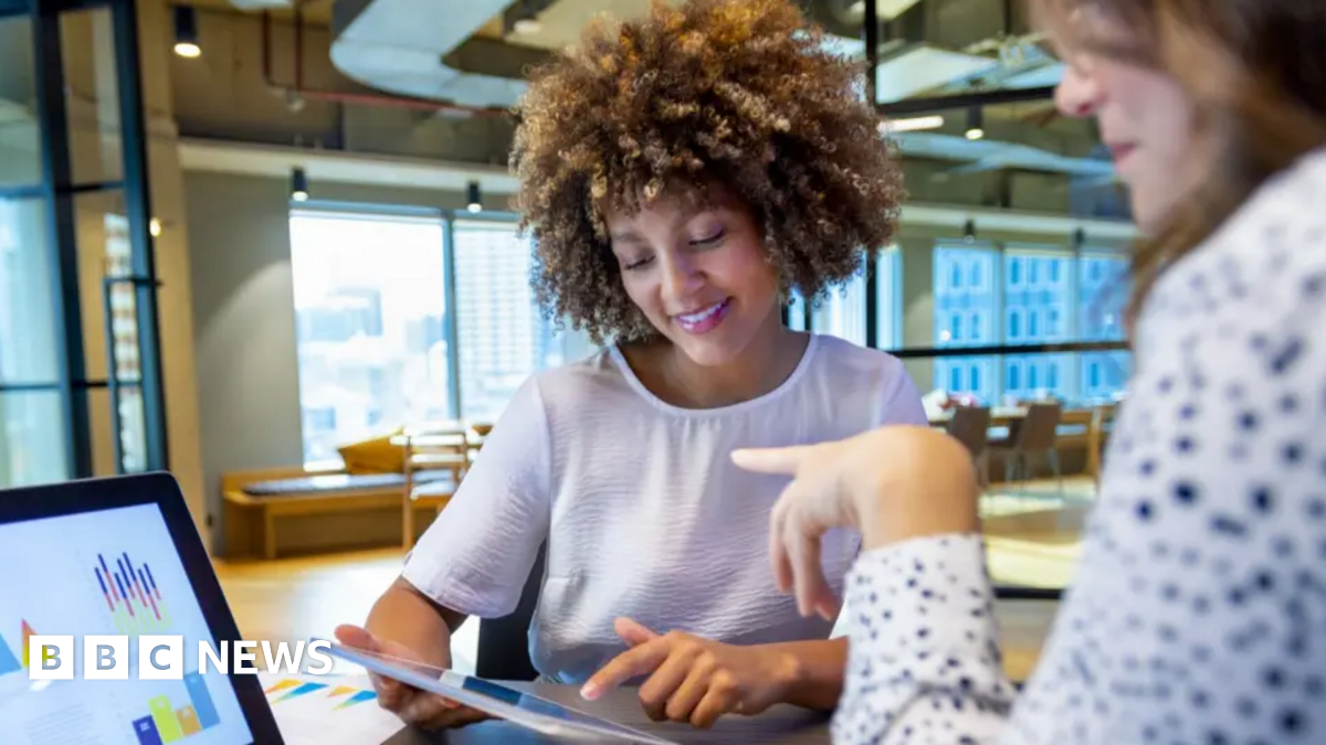 Two women sit opposite each other looking at a laptop screen and an iPad device. They are in an open-plan office with windows behind them as well as chairs and a desk.