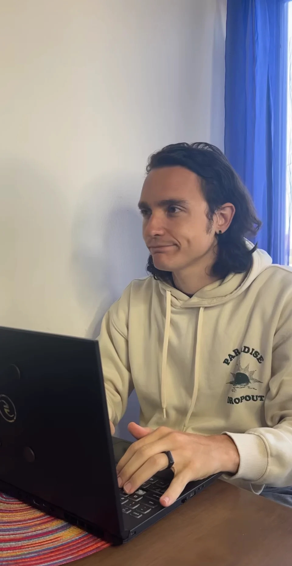 Person wearing a hoodie, sitting at a table, typing on a laptop