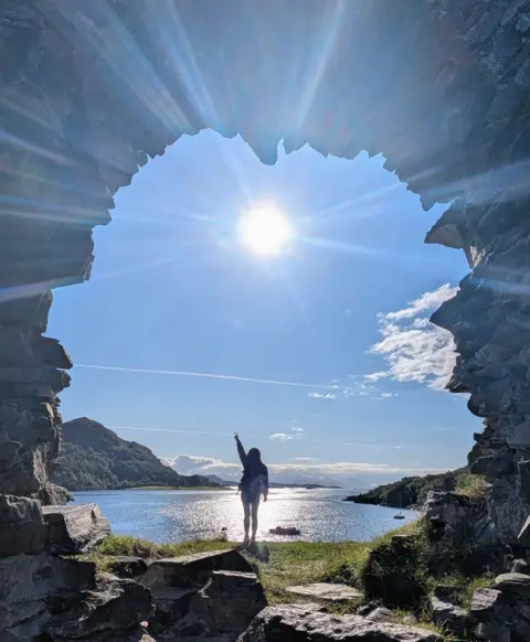 Tan Yi Shu We look through a stone archway to see a woman standing on a rock, the loch behind her, the sun shining on the water and in the sky, dead centre under the arch. 