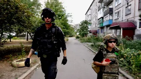 BBC News Two people - a tall man in black and a small woman in khaki camouflage gear, both wearing padded body warmers and dark sunglasses - walk down a residential street. Neither are smiling