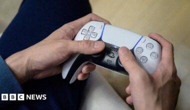 A gamer dressed in a navy blue top plays on the new Sony PlayStation 5 at his home. The photo is a close-up of his hands holding on to a white PS5 controller.