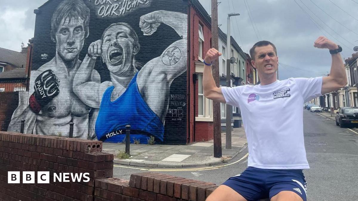 Joe Wild during his marathon in Liverpool, posing with his biceps flexed in front of a mural of martial arts fighters Paddy Pimblett and Molly McCann.