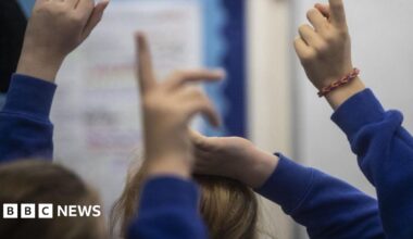 A generic image of unidentified primary school pupils holding their hands up while wearing blue sweatshirts.