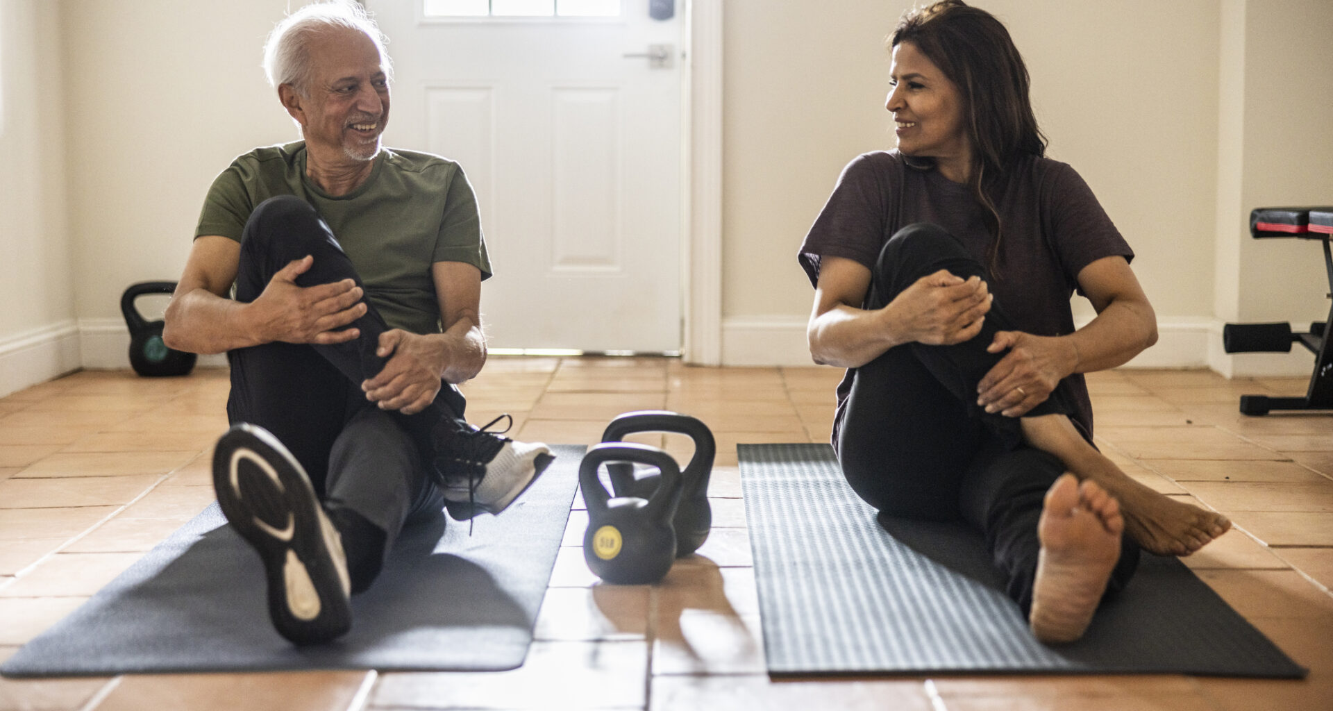 Senior man and woman sit on exercise mats performing a glute stretch, with kettlebells on the floor between them