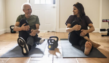 Senior man and woman sit on exercise mats performing a glute stretch, with kettlebells on the floor between them