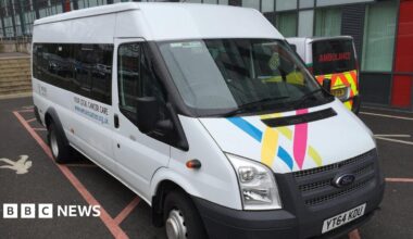 A white Ford Transit minibus with Wessex Cancer Support branding parked outside a hospital, with an ambulance beside it.