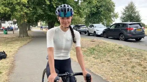 Katherine Barnes pictured on her road bike on a path surrounded by some grass and parked cars. She is wearing a cream coloured Lyrca cycling top and some black cycling shorts. She is also wearing a dark-blue helmet and has long brown hair in a French plait.