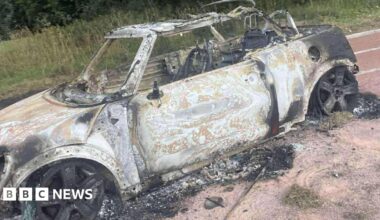 A burnt-out Mini car sitting on a cinder running track. The tyres have been completely destroyed. The wheel rims are fully blackened. The car has been stripped of all its paint and has lost its roof.