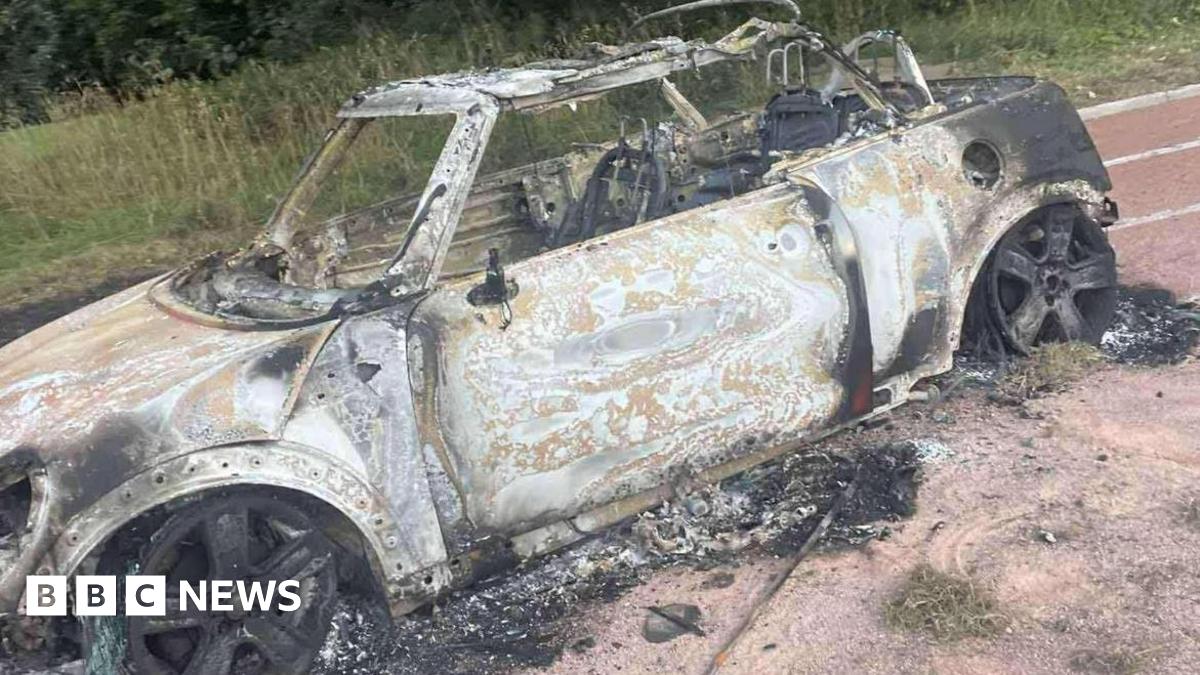 A burnt-out Mini car sitting on a cinder running track. The tyres have been completely destroyed. The wheel rims are fully blackened. The car has been stripped of all its paint and has lost its roof.