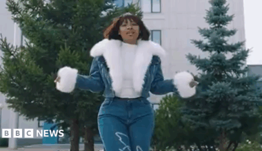 Cyan Boujee wearing jeans and a fur-lined jean jacket stands in front of two cedar trees and an office building. She is holding her arms out as she addresses the camera.