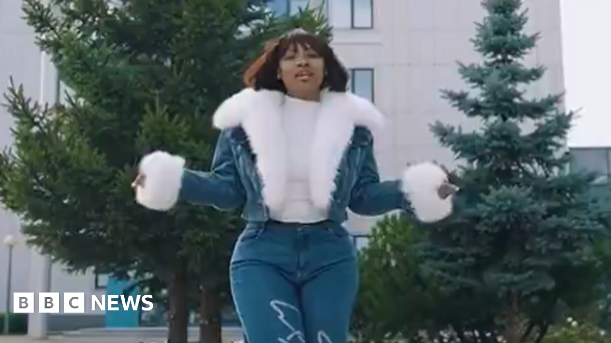 Cyan Boujee wearing jeans and a fur-lined jean jacket stands in front of two cedar trees and an office building. She is holding her arms out as she addresses the camera.