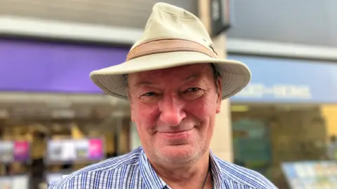 Greg is wearing a beige hat and blue and white checked shirt