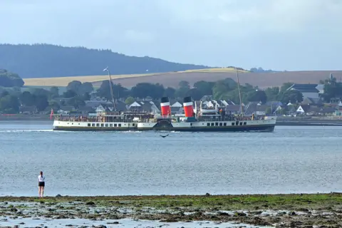 Eric Niven A shot of the red two-funnelled Waverly paddle steamer making its way along the River Tay. A woman is on the edge of the shore taking a picture.