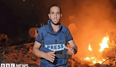 Anas al-Sharif is wearing a blue flak jacket with 'PRESS' written across the front, and he's standing in front of burning debris.