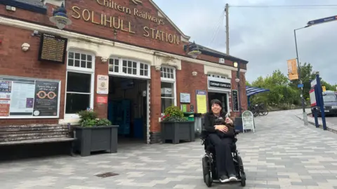 A woman in a wheel chair sitting infant of a railway station. She is wearing all black and has black hair. The station is red brick. 