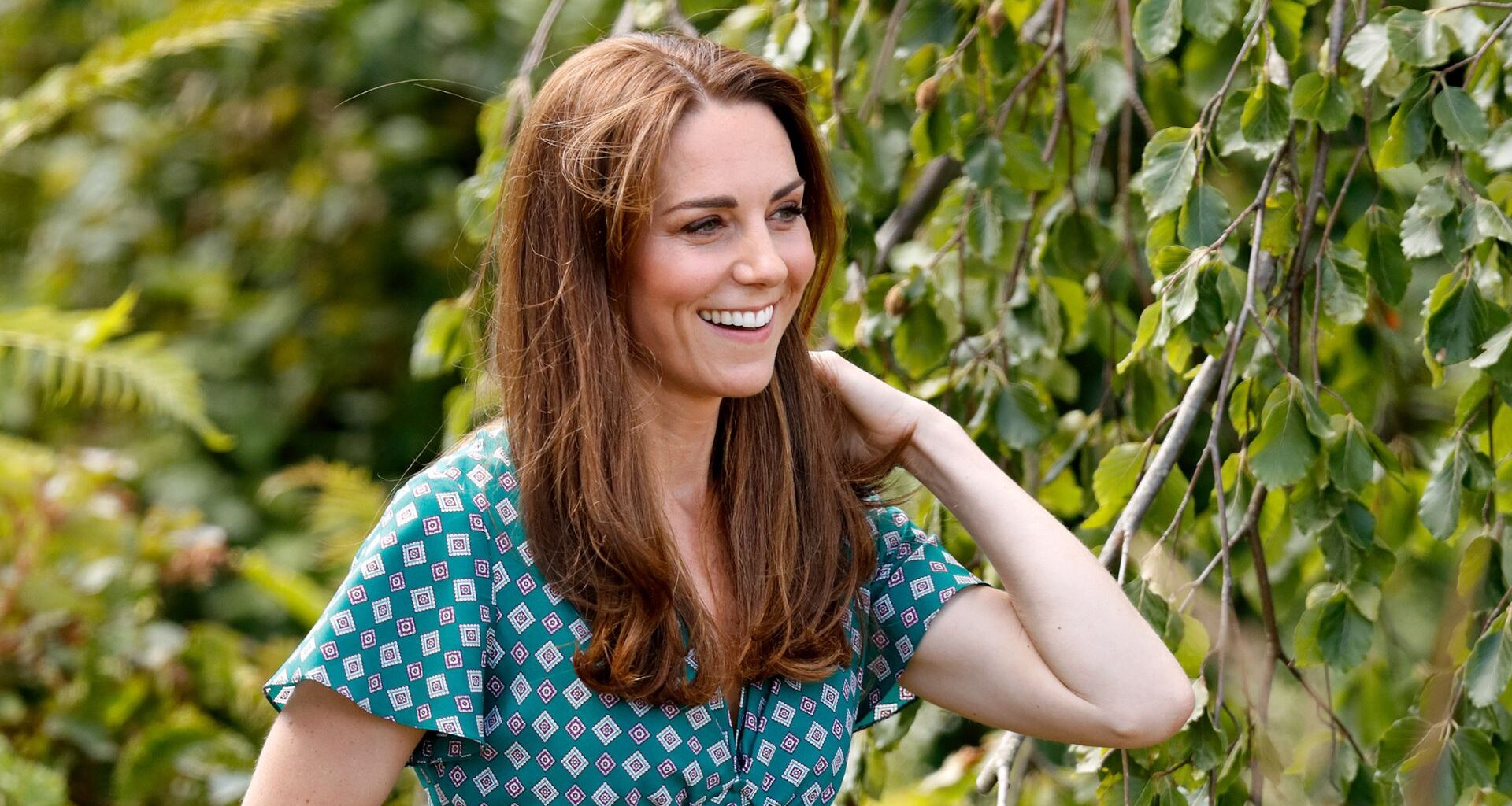 Kate Middleton smiling as she stands outdoors near some trees and plants
