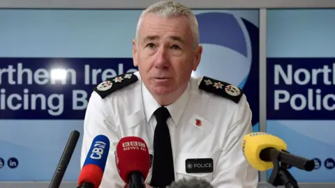 Getty Images A man sits at a table with several mics belonging to media organisations in front of him. Lapels are on his shirt. The word police is printed on his shirt on a badge. 