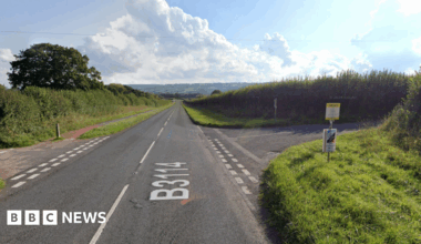 A rural road with grass verges and junctions either side of the road.
