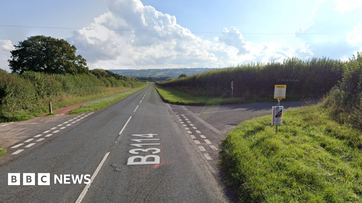 A rural road with grass verges and junctions either side of the road.