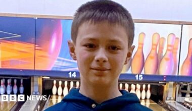 A young boy, Leo Ross, has dark brown hair and is wearing a navy hoodie. He is standing in front of 10-pin bowling alley lanes. Above the lanes are images of multi-coloured bowling skittles that are yellow, pink and blue.