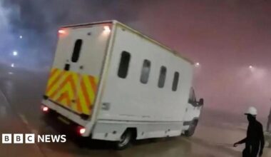 A screenshot from a video uploaded by Palestine Action. It shows a white van with curved rectangular windows and yellow and orange hi-vis decals on the back. It is night time and there is a lot of smoke in the air which is being lit up by the van's red brake lights. On the right there is a figure wearing all black and a white hard hat.