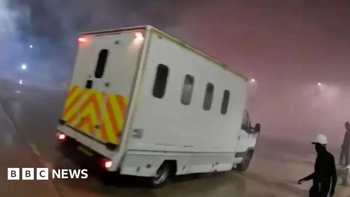 A screenshot from a video uploaded by Palestine Action. It shows a white van with curved rectangular windows and yellow and orange hi-vis decals on the back. It is night time and there is a lot of smoke in the air which is being lit up by the van's red brake lights. On the right there is a figure wearing all black and a white hard hat.