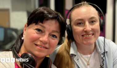 Nataliia Revkova and Nadiia Hoian smile at the camera. They are sat next to each other in a radio studio. Nataliia has dark hair that is tied up and she has a fringe. Headphones rest around her neck. Nadiia has long blonde hair that is tied up and she wears her headphones.