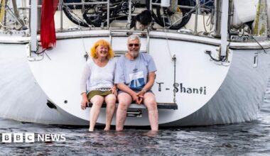 Debbie and Stephen sitting on the back of their yacht with their feet in the water. The word "T-Shanti" is printed on the back of the white boat. Both are smiling at the camera.
