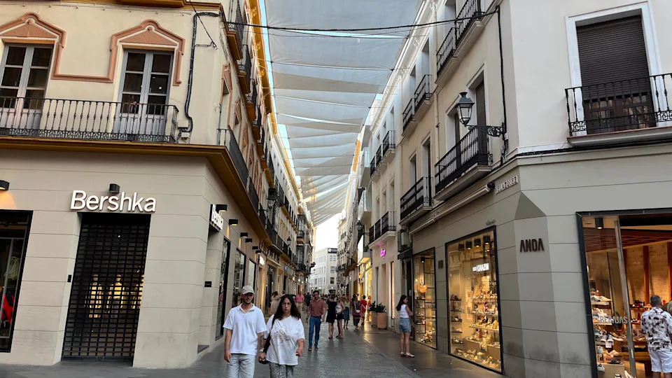 A crowd walks down a street with awnings pulled across roofs of the buildings creating shade