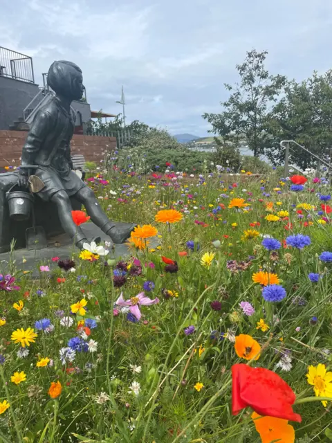 Marjory Robertson A bronze sculpture of a young girl sitting down is surrounded by a sea of wildflowers in bright orange, purple, pink and red with a row of trees in the distance and beyond that, the Firth of Clyde.