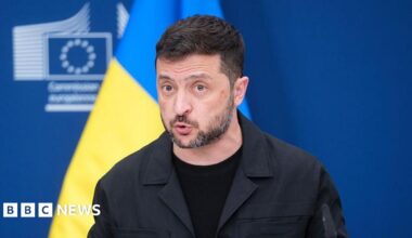 Ukrainian President Zelensky talks to the press while standing in front of a blue and yellow Ukrainian flag.