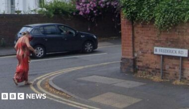 A woman dressed in a long red dress is crossing Frederick Road at its junction with St James Road, on which a blue car is being driven. The sign for Frederick Road is showing on the right of the shot.