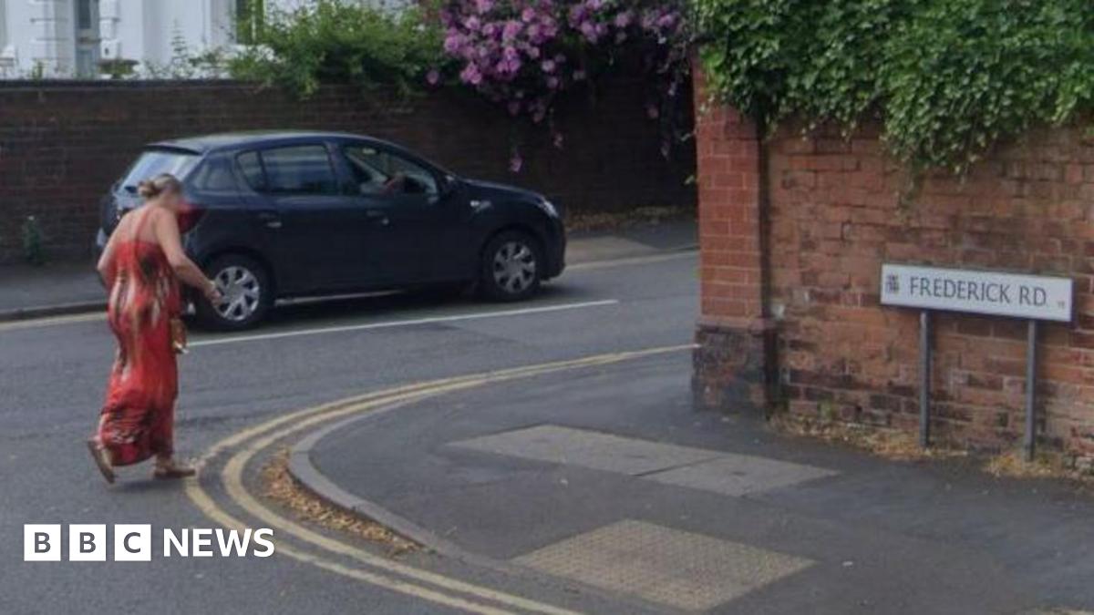 A woman dressed in a long red dress is crossing Frederick Road at its junction with St James Road, on which a blue car is being driven. The sign for Frederick Road is showing on the right of the shot.