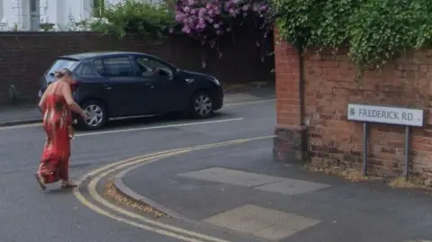 Google A woman dressed in a long red dress is crossing Frederick Road at its junction with St James Road, on which a blue car is being driven. The sign for Frederick Road is showing on the right of the shot. 
