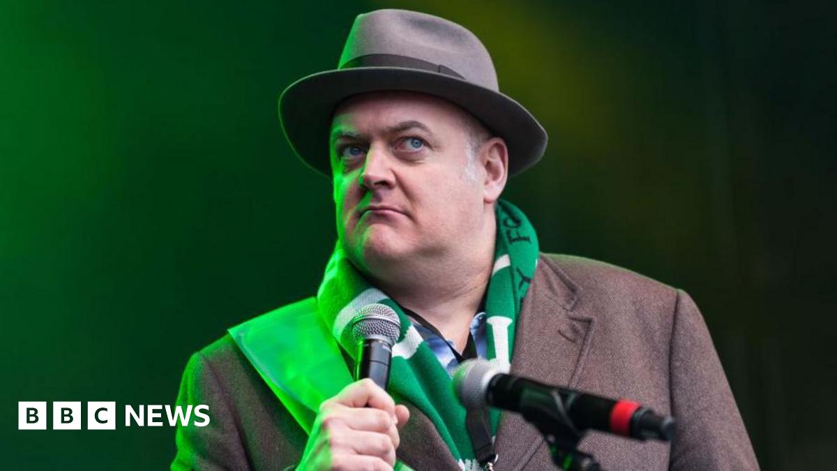 Dara O'Briain on stage holding a microphone. He is wearing a brown hat and overcoat and a green and white striped scaft.