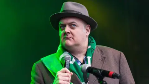 Getty Images Dara O'Briain on stage holding a microphone. He is wearing a brown hat and overcoat and a green and white striped scaft.