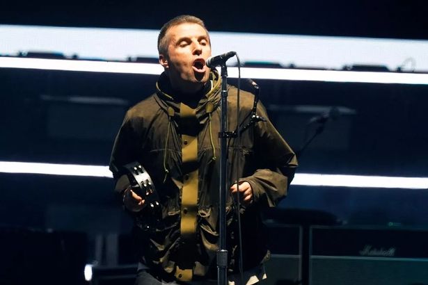 An image of a short-haired man wearing a parka jacket, singing into a microphone while holding a tambourine.