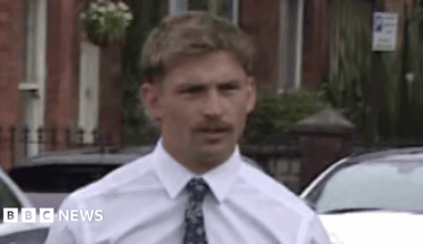 Kavan Keenan is a young man with short blonde/ brown hair and a moustache. He is walking towards the gates into the court as the picture is taken. He is wearing a white long sleeve shirt and a black patterned tie. Behind him is a silver car and two white cars parked along the street in front of red brick terrace houses.