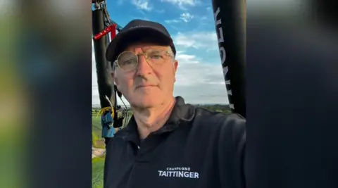 Phoenix Balloons Pete Dalby. He is wearing a black hat and reading glasses. He is pictured in a hot air balloon. He is looking at the camera and squinting in the sun.