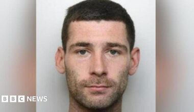 Michael Trainer's mugshot, where he is staring at the camera with a serious expression and has dark brown hair and dark stubble. He is stood in front of a light grey background.