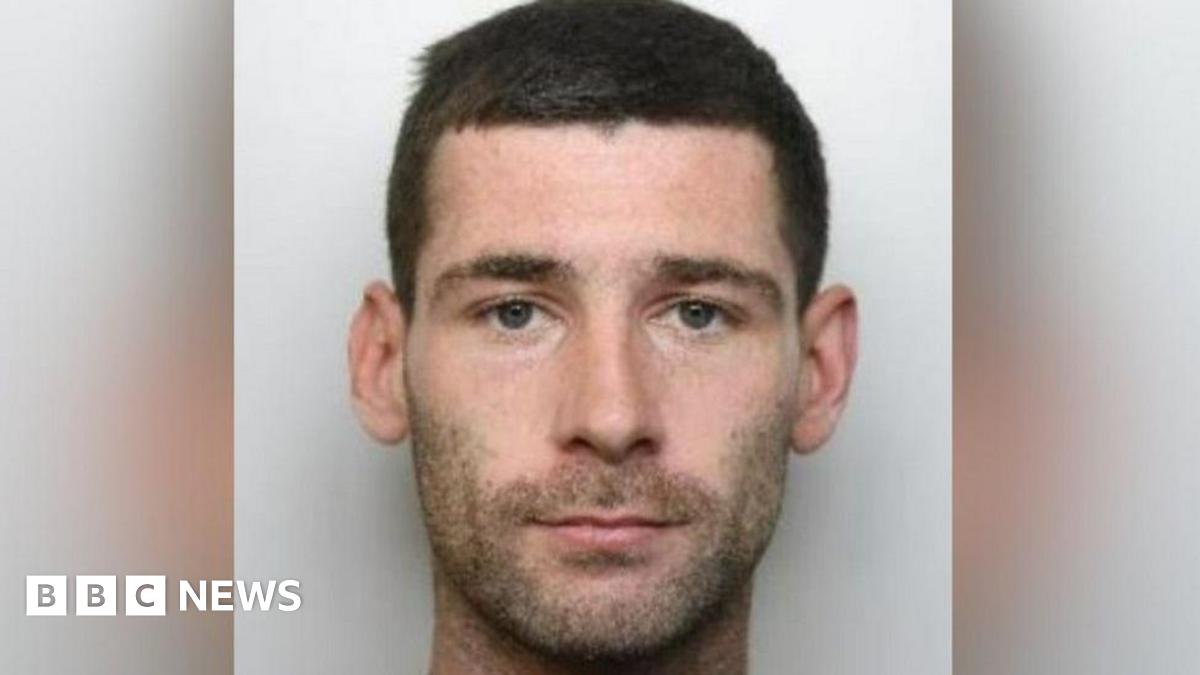 Michael Trainer's mugshot, where he is staring at the camera with a serious expression and has dark brown hair and dark stubble. He is stood in front of a light grey background.