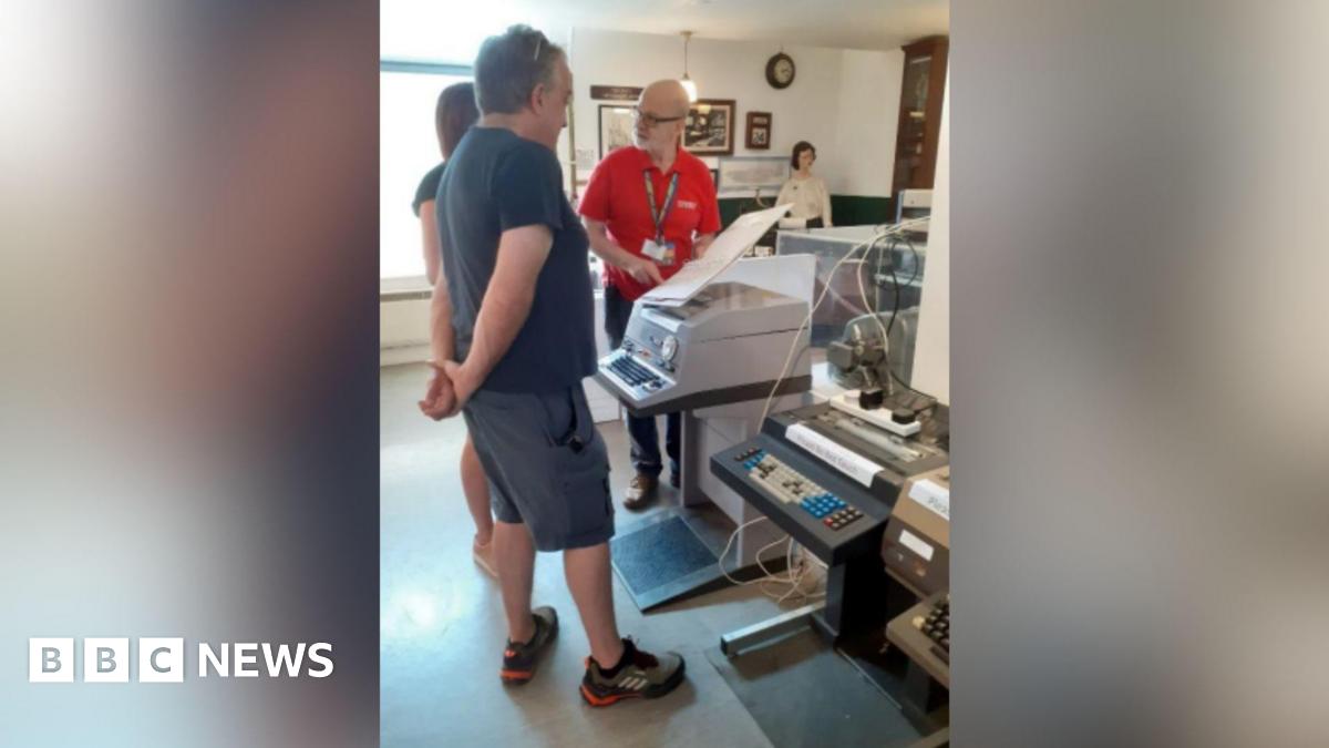 A man in a red polo shirt demonstrating to two people how a large grey Telex machine works