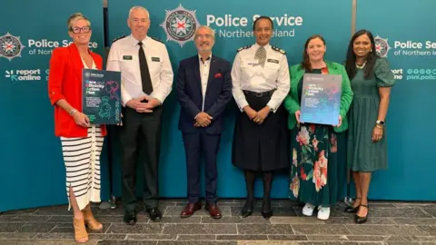 PSNI Jon Boutcher, along with five other people, standing in front of a large PSNI poster. All six are smiling at the camera, and the woman on the far left is holding a sign displaying the name of the strategy. 