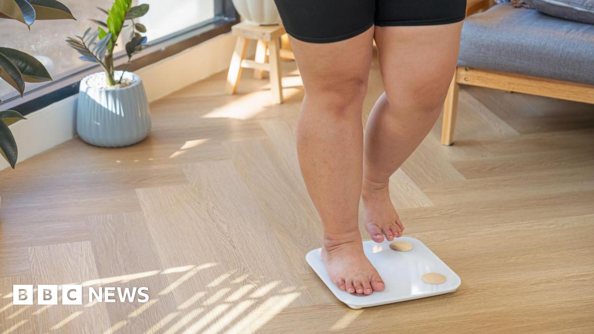 Waist down shot of a woman standing on weighing scales, wearing black gym shorts. She has bare legs and feet.  She is in a bright living room which has a wooden floor and potted plants in the background.