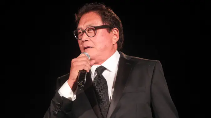 Robert Kiyosaki in a suit talking at an event, holding a microphone