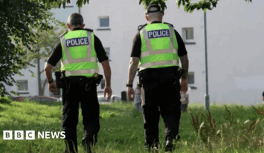 Two police officers wearing hi-viz vests with police written on the back over dark clothing walk on a patch of grass in front of a light coloured block of flats with small windows on a sunny day.