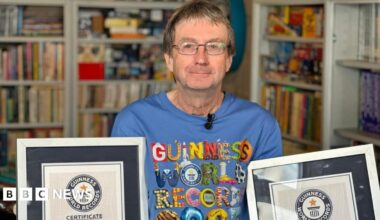 A man is standing in a room with bookshelves around him filled with Guinness World Record books. He is wearing a blue T-shirt with the Guinness World Record logo on it. He is holding up two certificates.