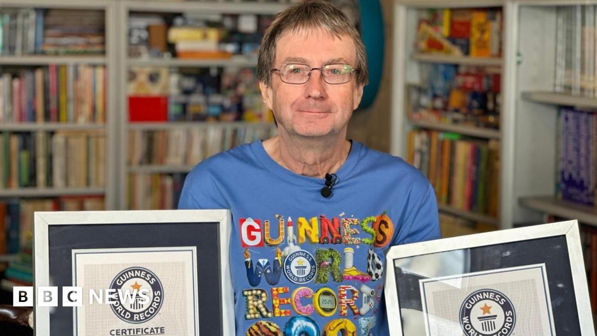 A man is standing in a room with bookshelves around him filled with Guinness World Record books. He is wearing a blue T-shirt with the Guinness World Record logo on it. He is holding up two certificates.